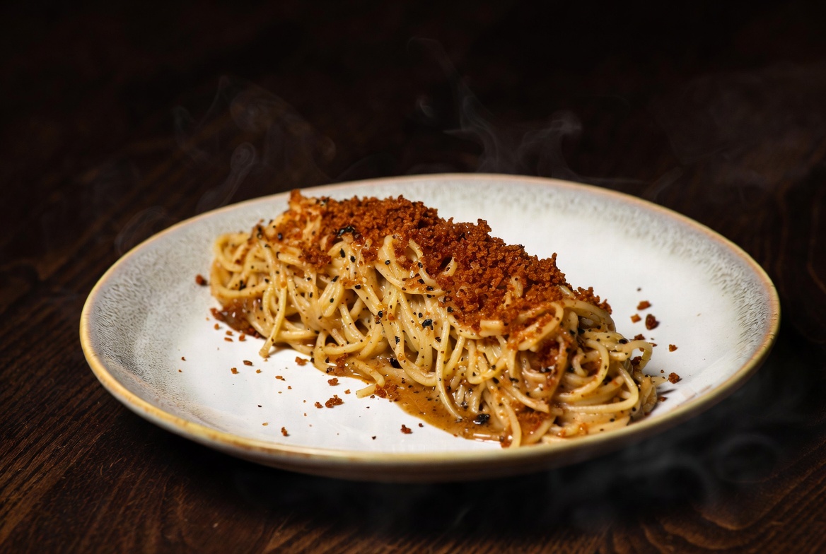 Wide editorial frame of a plated pasta dish with linen, ceramic, and amber room light.